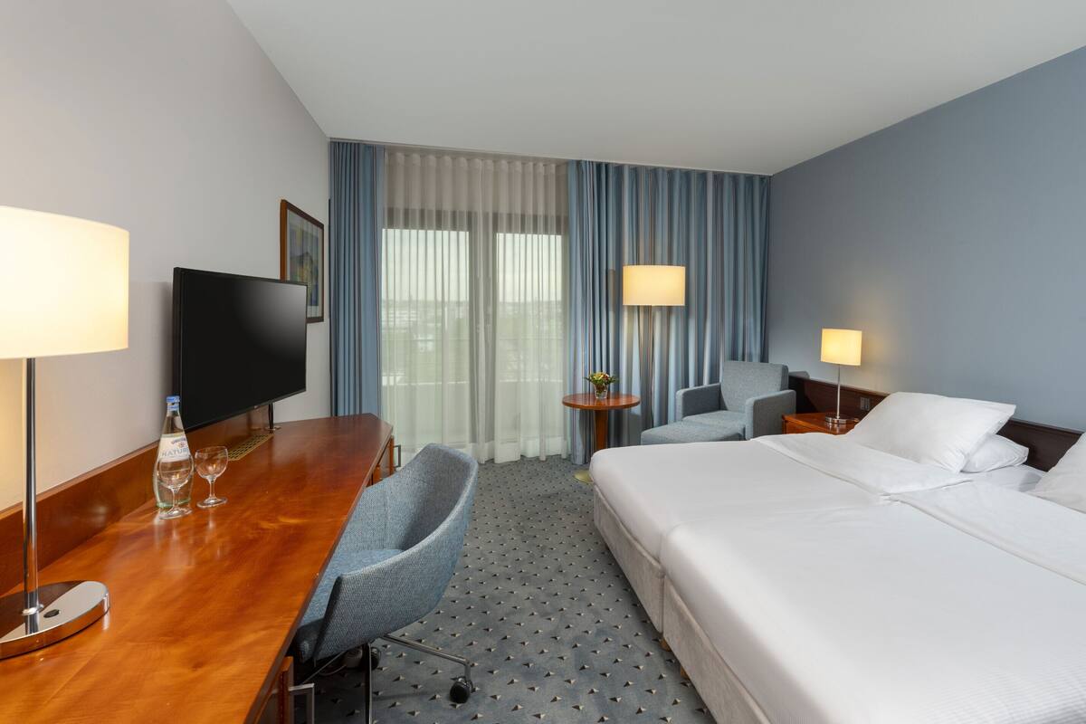 Habitación de hotel con cama, escritorio con televisor, silla, dos mesitas de noche con lámparas y un ventanal con cortinas.