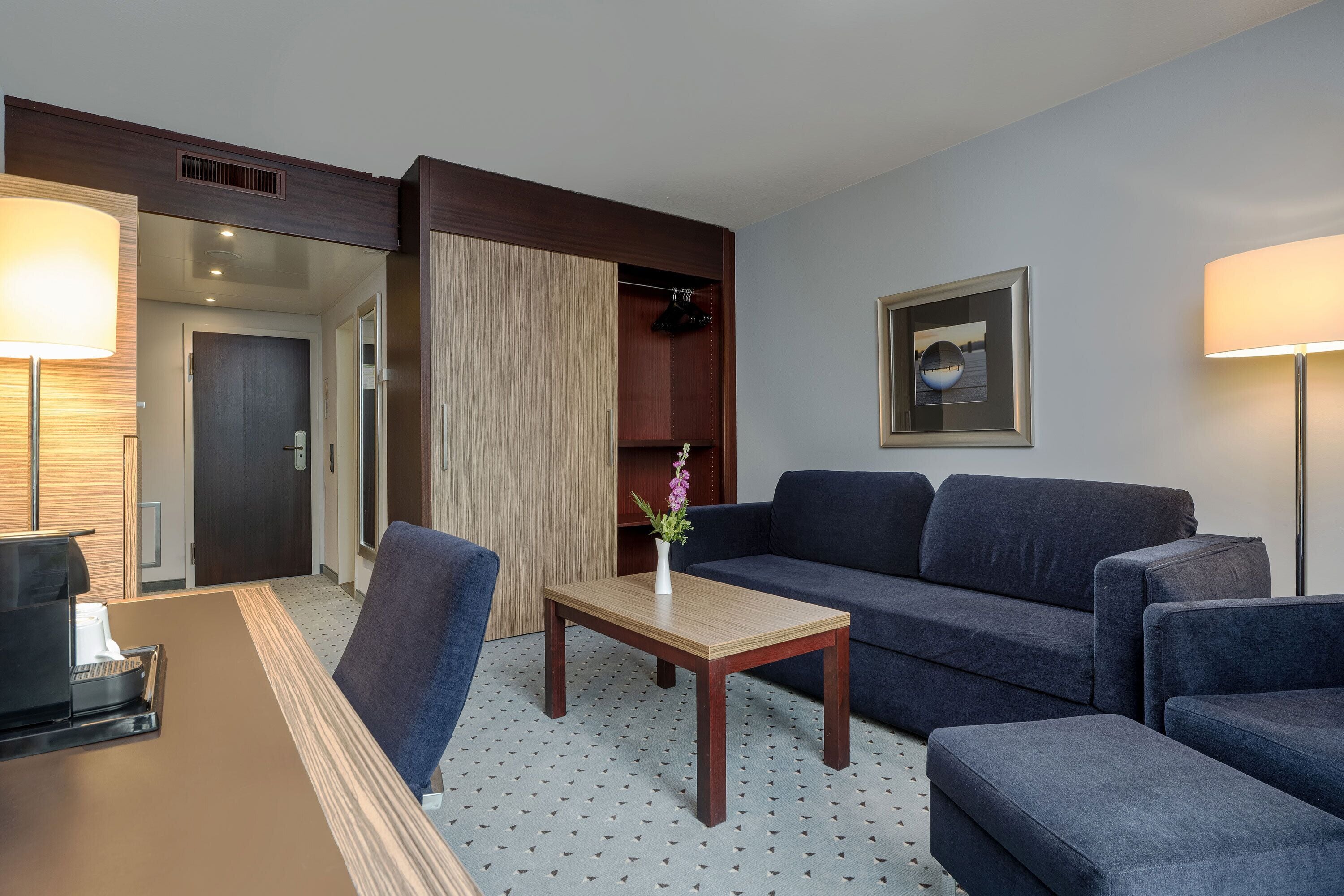 A hotel room with a sofa set, a wooden coffee table, a desk with a lamp, and a framed picture on the wall.