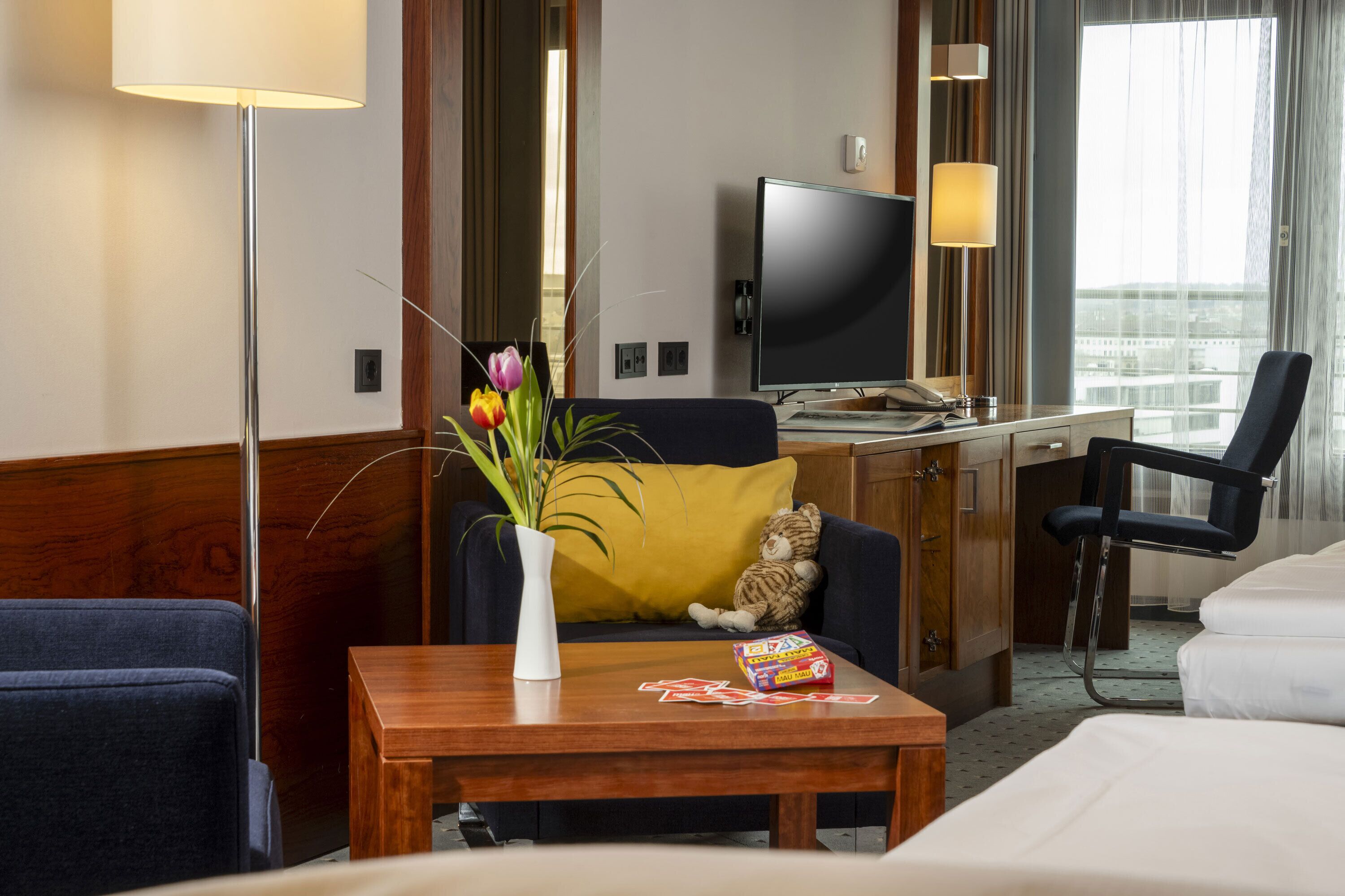 Ein Hotelzimmer mit einem Bett, einem Schreibtisch, einem Stuhl, einem Fernseher, einer Lampe und einer Blumenvase.