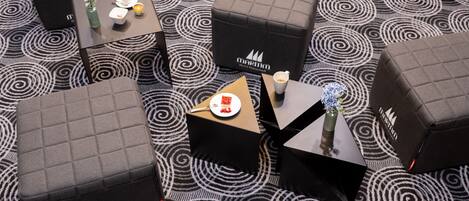 Una habitación de hotel con alfombra estampada, varios otomanes, una mesita con una taza y platillo, y un florero con flores.