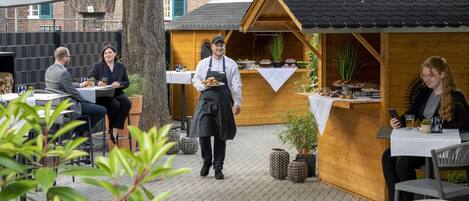 An outdoor dining area with tables and chairs, a wooden structure with a tiled roof, and potted plants.