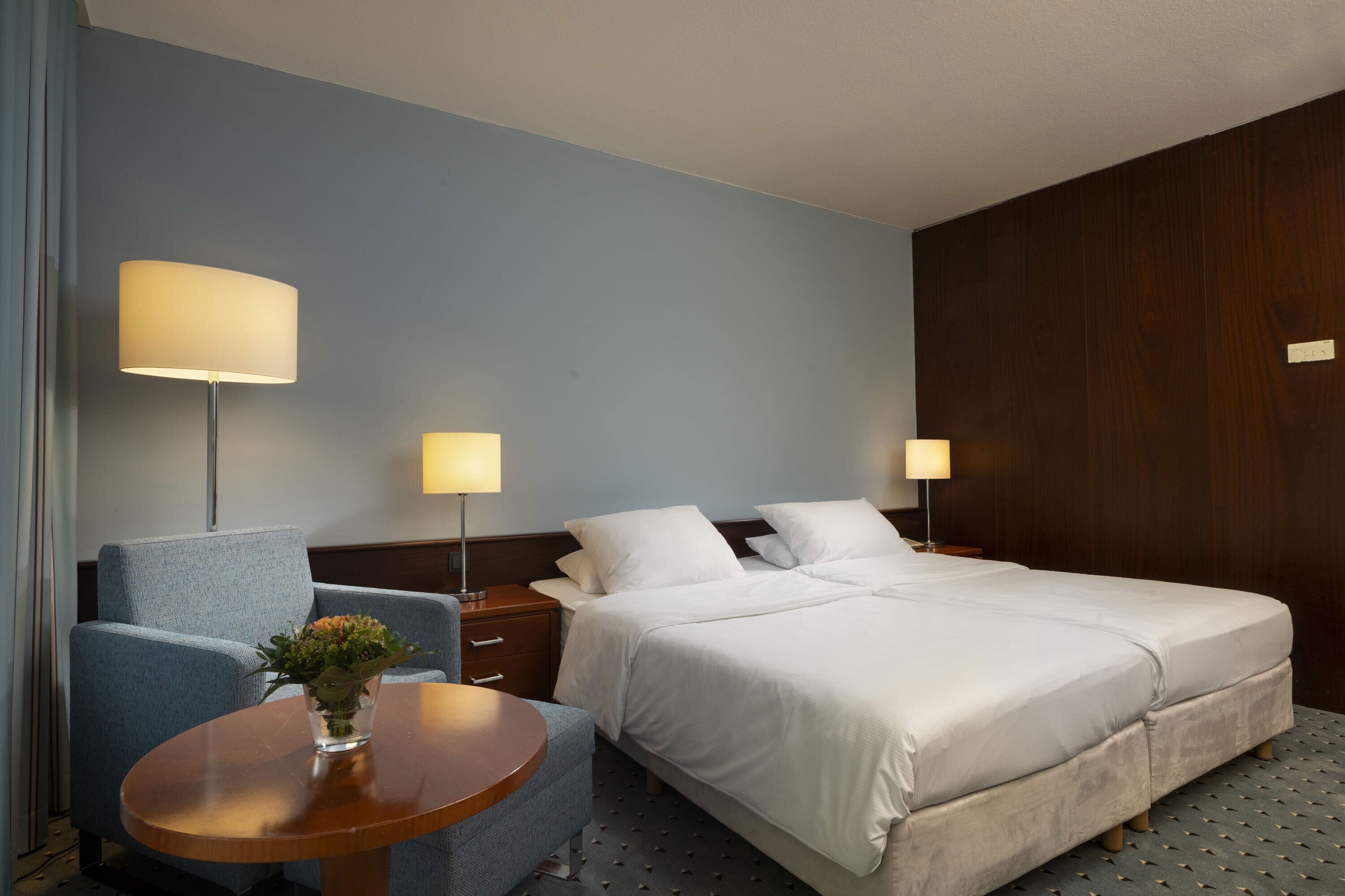 Een hotelkamer met een tweepersoonsbed, een stoel, een klein tafeltje en twee bedlampjes.