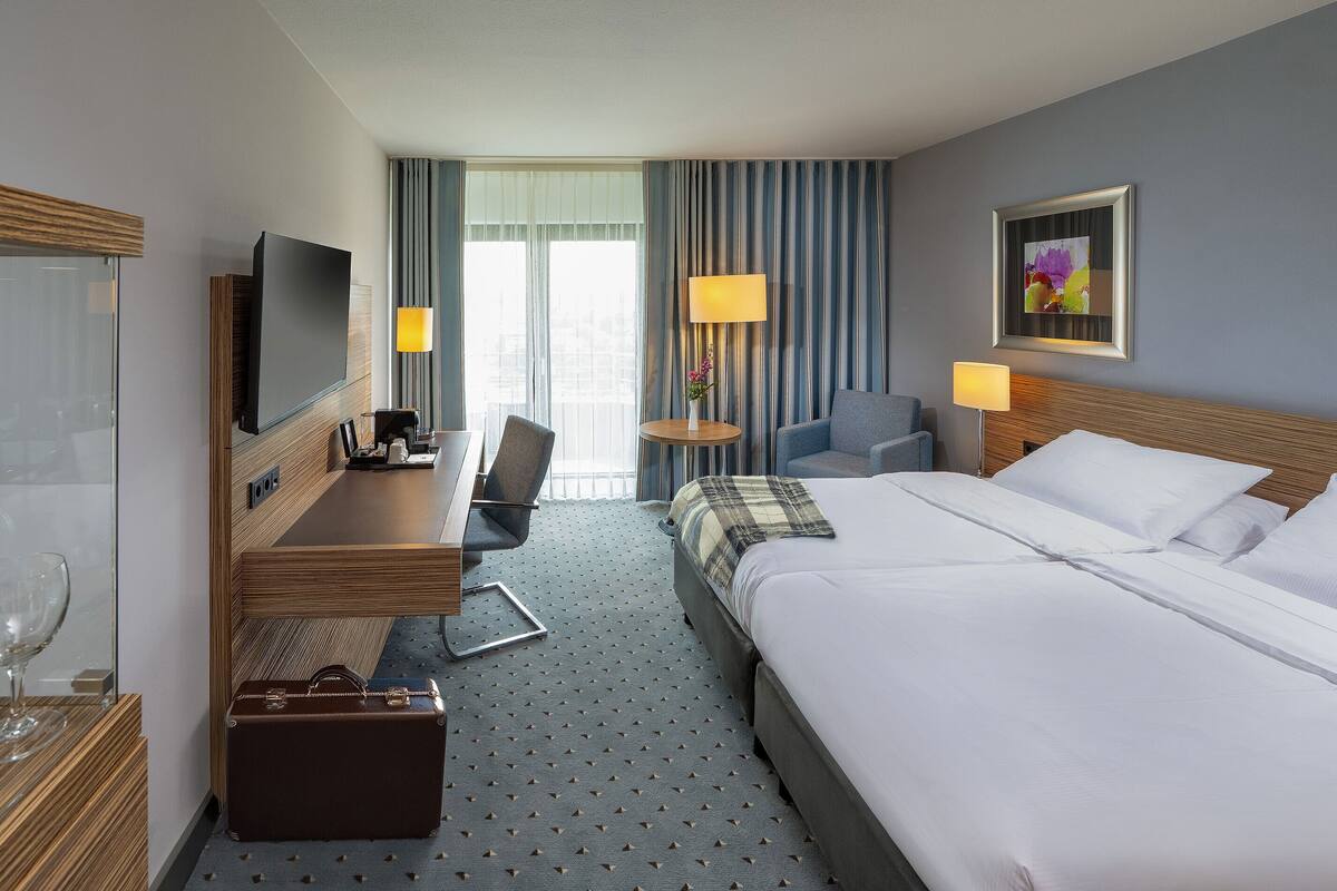 Habitación de hotel con cama, escritorio con televisor de pantalla plana, una silla, una mesita con lámpara y maleta en el suelo.