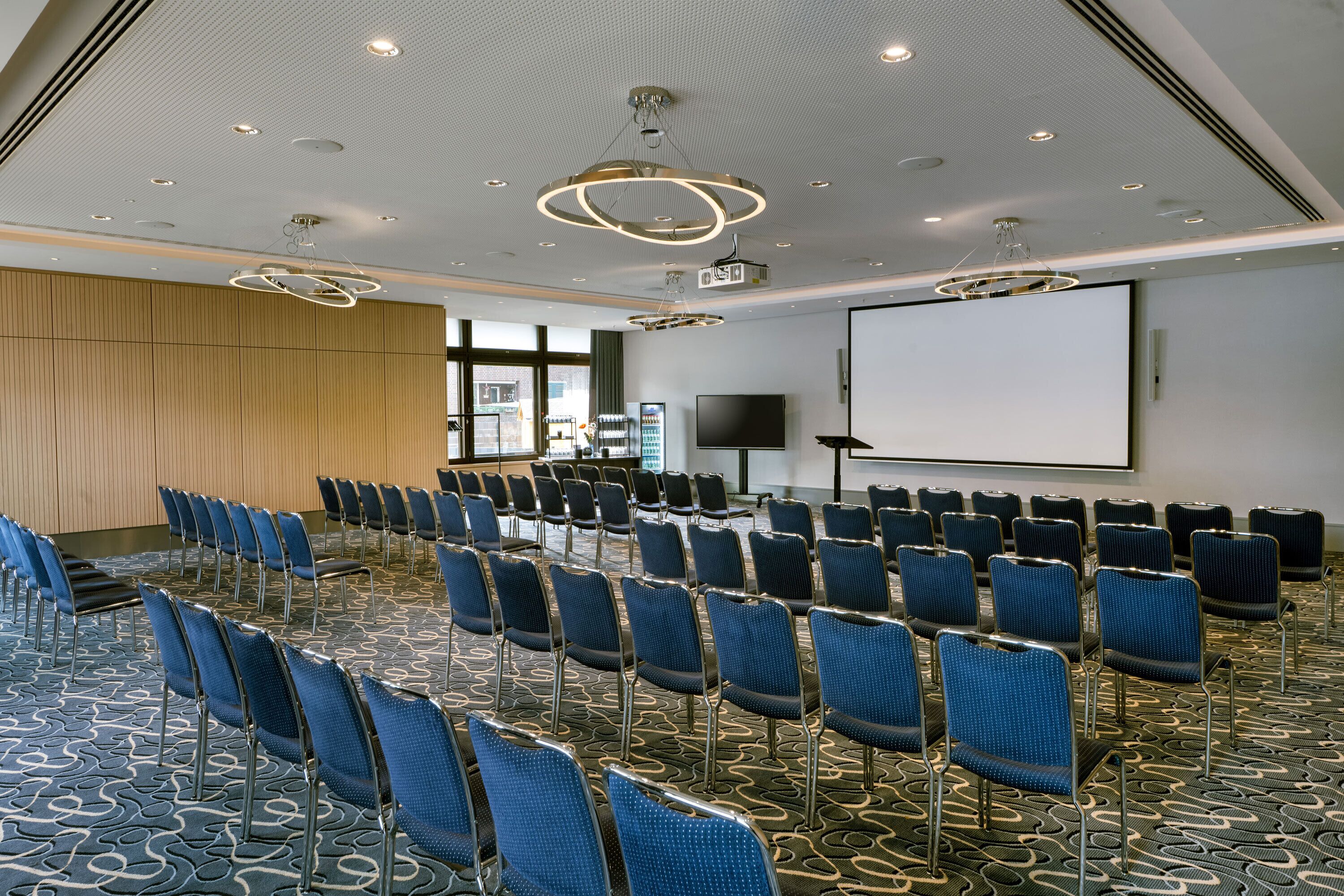 Une salle de conférence avec des rangées de chaises bleues, un grand écran et un projecteur.