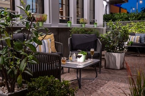 An outdoor seating area with cushioned chairs and tables, surrounded by potted plants and a stone floor.