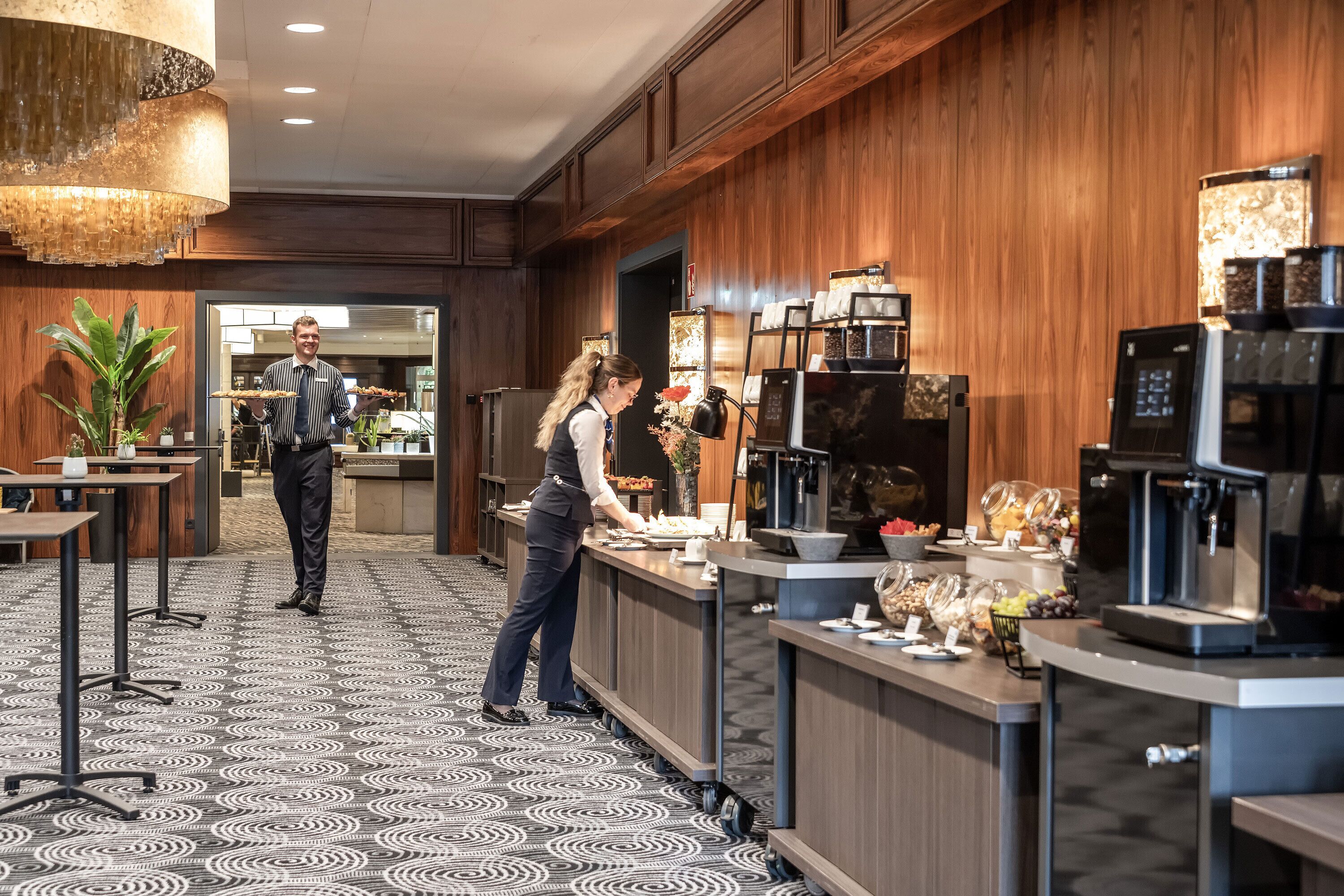 Een ontbijtruimte van een hotel met een lange balie, koffieautomaten en een buffet.