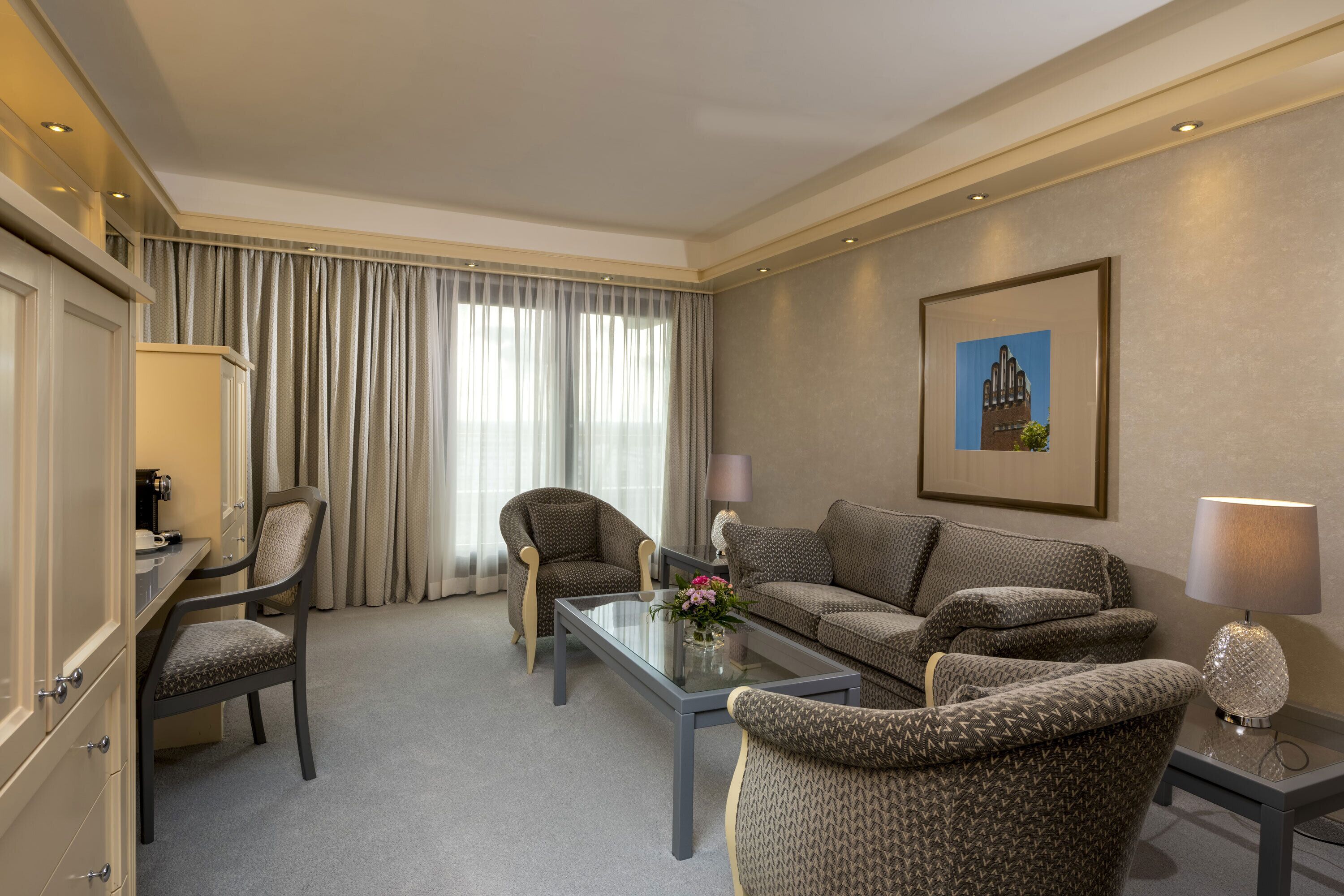 Habitación de hotel con sofá, dos sillones, una mesita de centro, escritorio con lámpara y un cuadro en la pared.