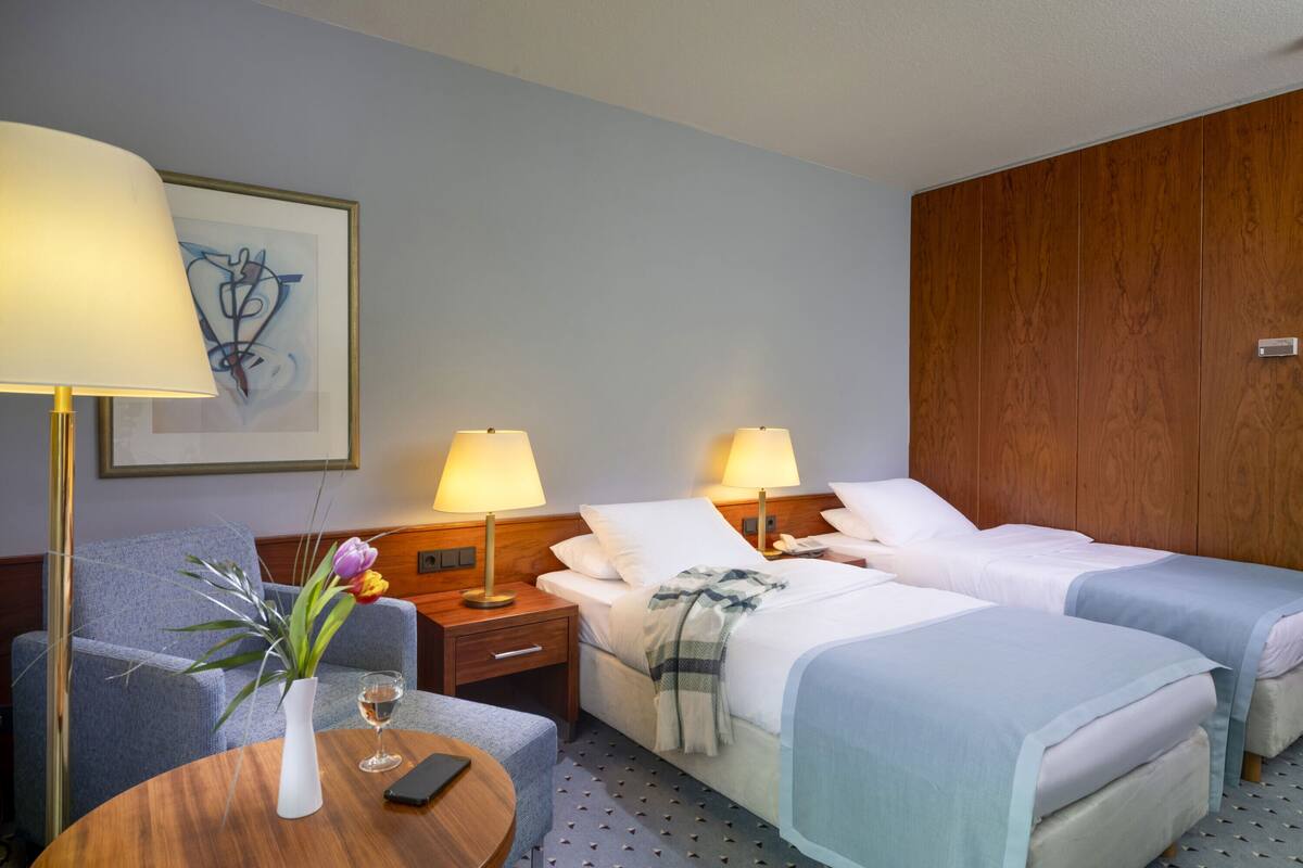 Habitación de hotel con dos camas, cabecero de madera, una mesa redonda con un florero y una copa de vino, y una obra de arte enmarcada en la pared.