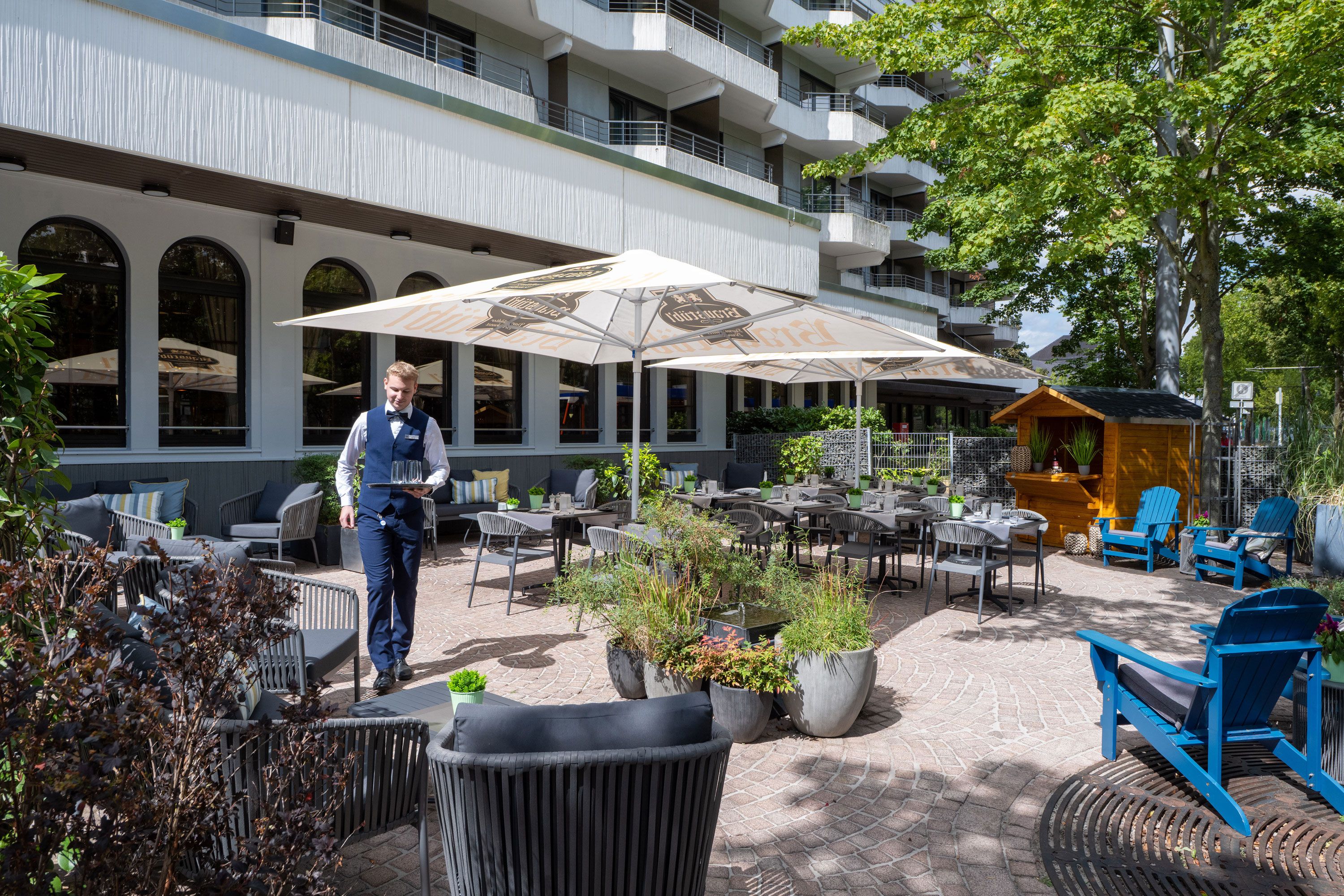 Un patio al aire libre con mesas y sillas, un camarero llevando una bandeja y un edificio con balcones al fondo.
