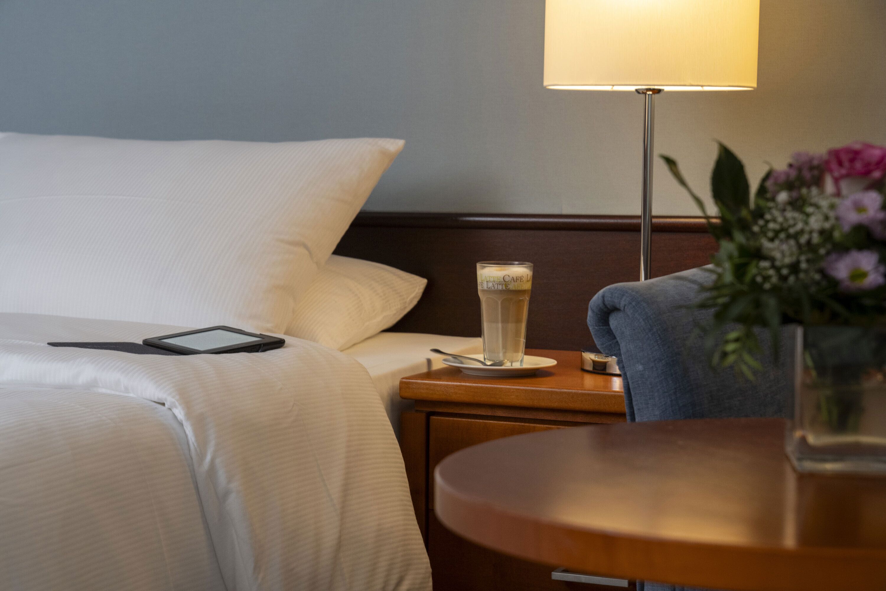 Ein ordentlich bezogenes Bett mit weißen Laken, ein Nachttisch mit Lampe, einem Glas Wasser und einem Tablet.