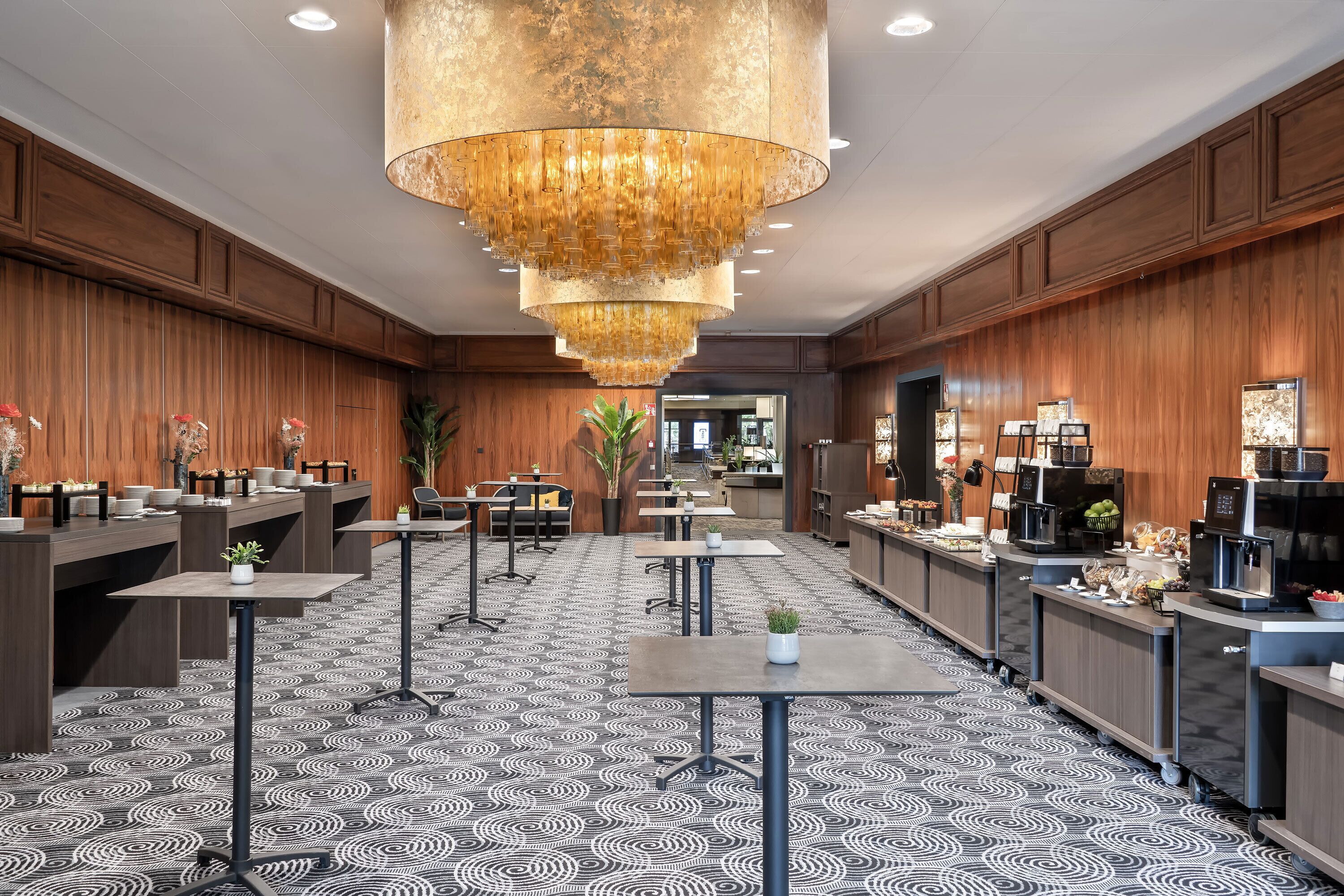A hotel lobby with a patterned floor, wooden paneling, a large chandelier, and a reception area with a coffee machine and counter.