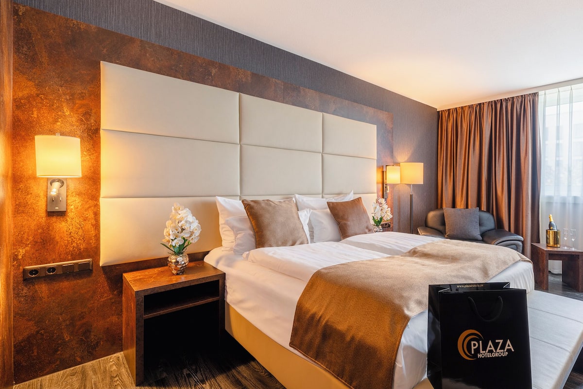 A hotel room with a large bed, two bedside tables, a desk, a chair, and a large window with curtains.