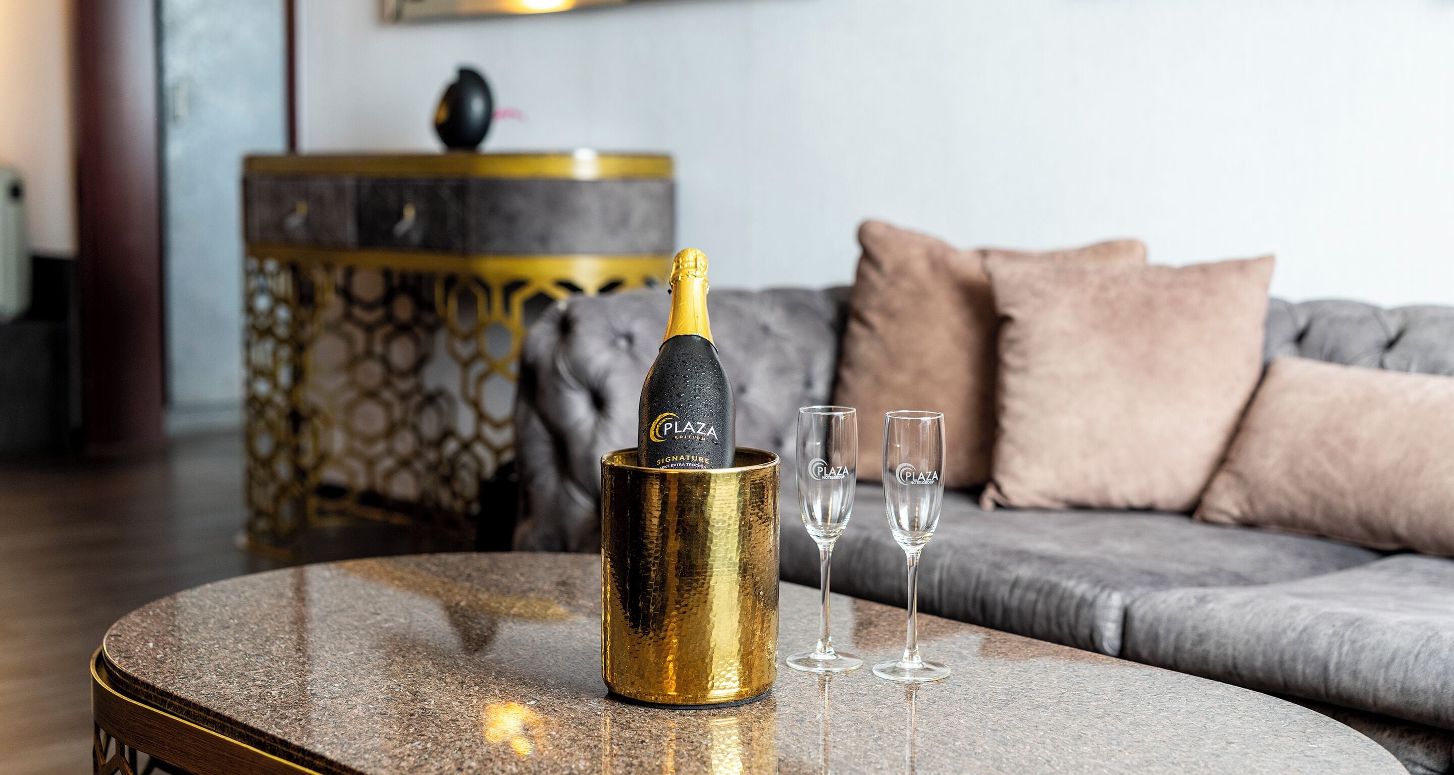 A bottle of champagne in a gold holder on a round table, two champagne flutes, a grey sofa with cushions, and a decorative side table with a geometric pattern.