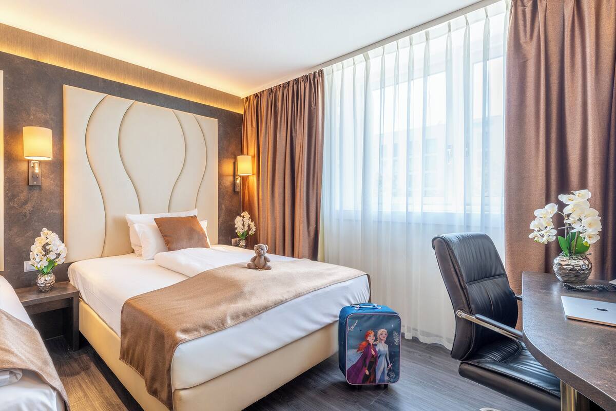 A hotel room with two beds, a desk, a chair, a suitcase, and a vase with flowers.