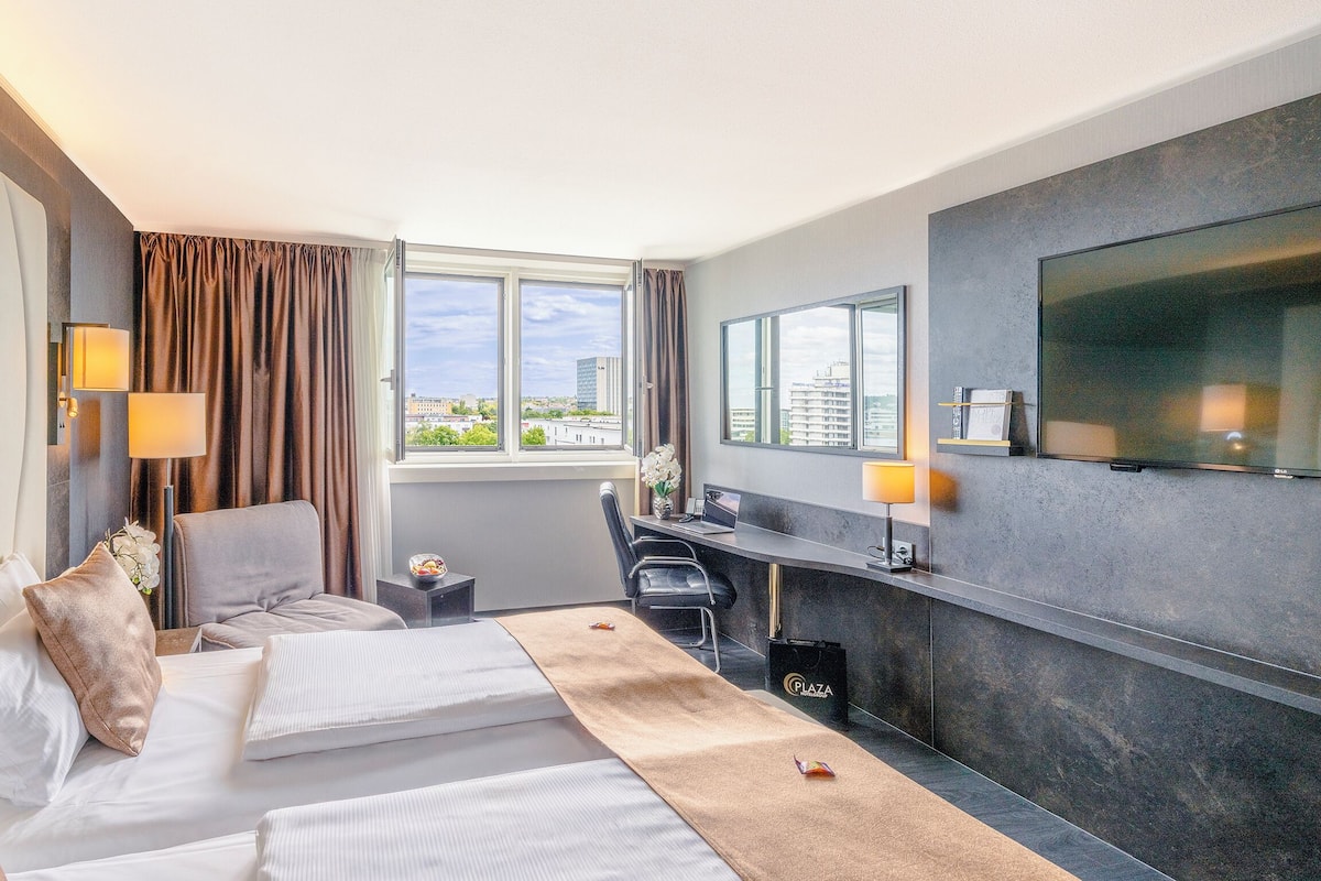 A hotel room with a bed, a desk, a chair, a TV, and a window with a city view.