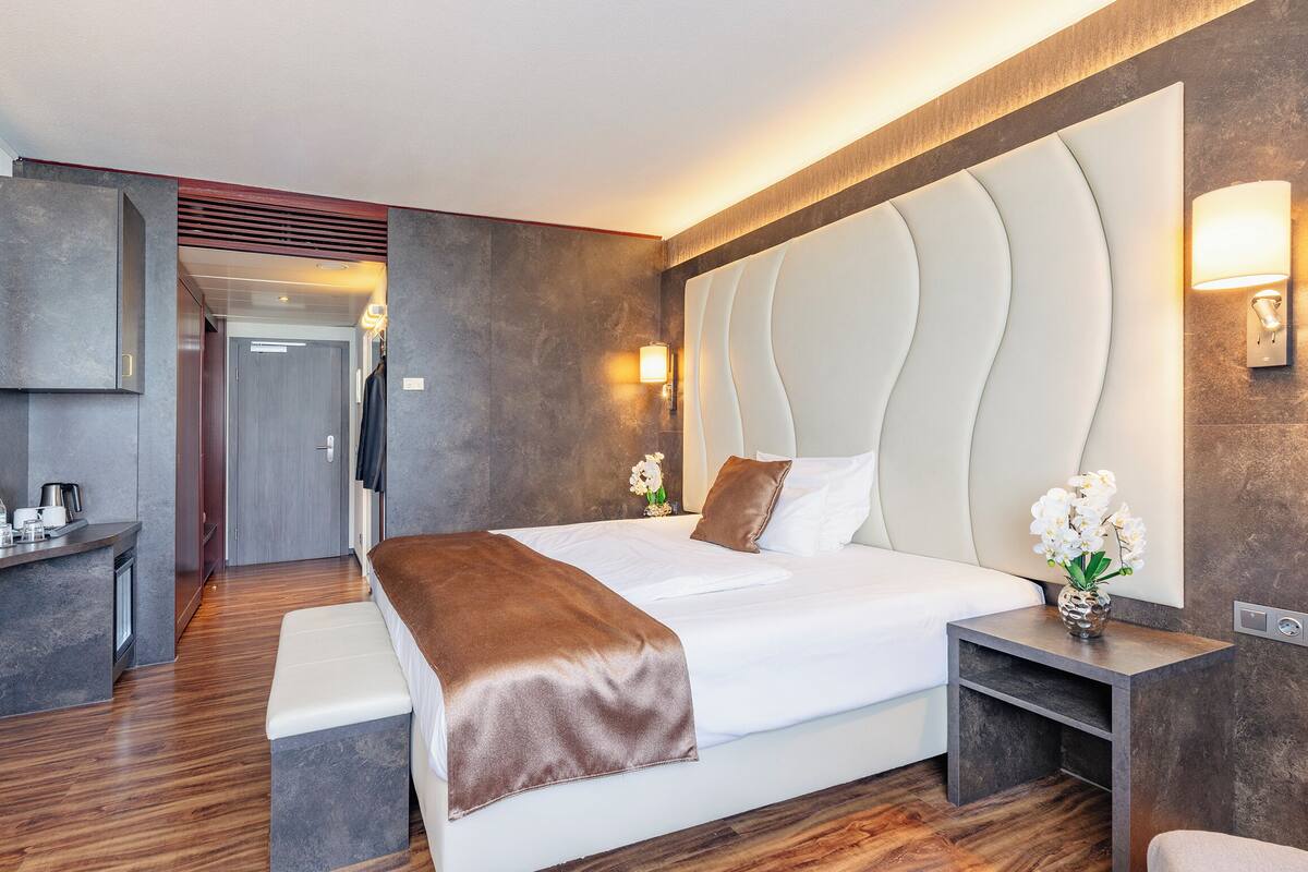 A modern hotel room with a large bed, a bedside table with a lamp, a mirror, and a small desk with a coffee maker.
