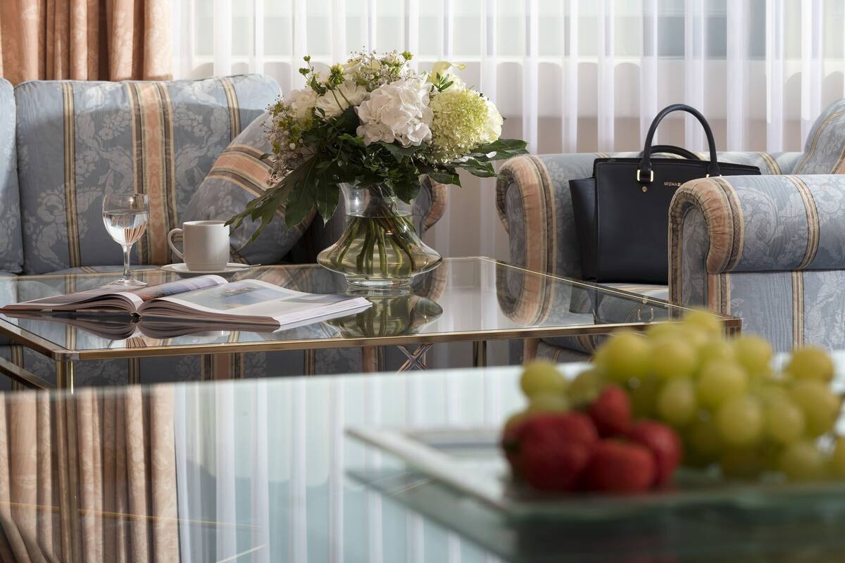 A living room with a sofa, a glass coffee table, a vase with flowers, a book, a cup, a handbag, and a bowl of fruit.
