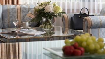 A living room with a sofa, a glass coffee table, a vase with flowers, a book, a cup, a handbag, and a bowl of fruit.