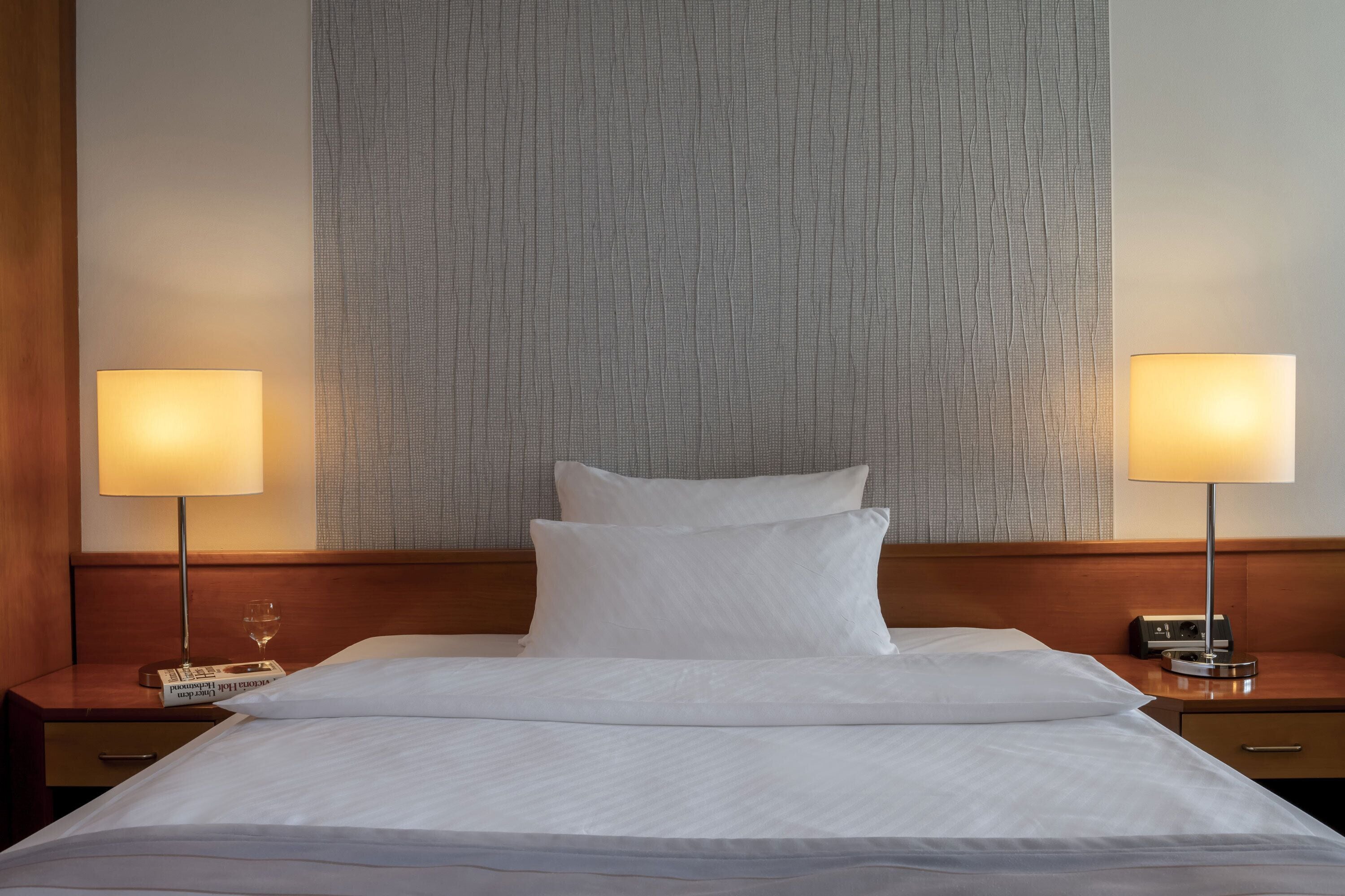 A neatly made bed with white linens, two bedside tables with lamps, a wooden headboard, and a textured wall behind the bed.