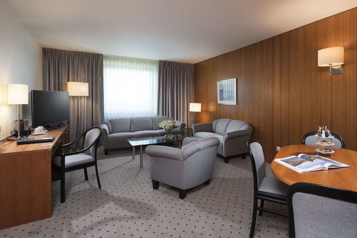 A hotel room with a wooden panel wall, a sofa, two armchairs, a dining table with chairs, a TV, and lamps.