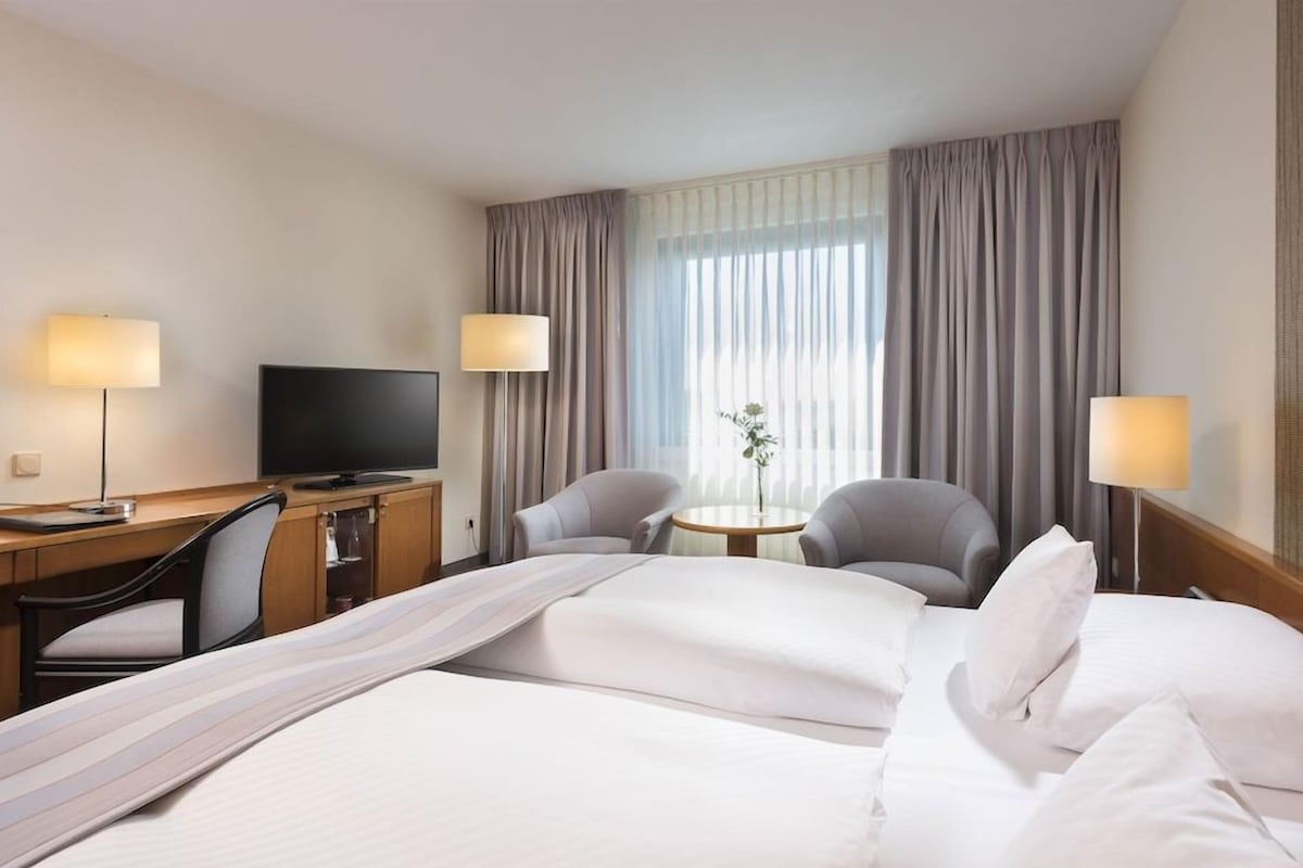 Una habitación de hotel con una cama, dos sillas, un escritorio, un televisor y una mesita de noche con un florero.