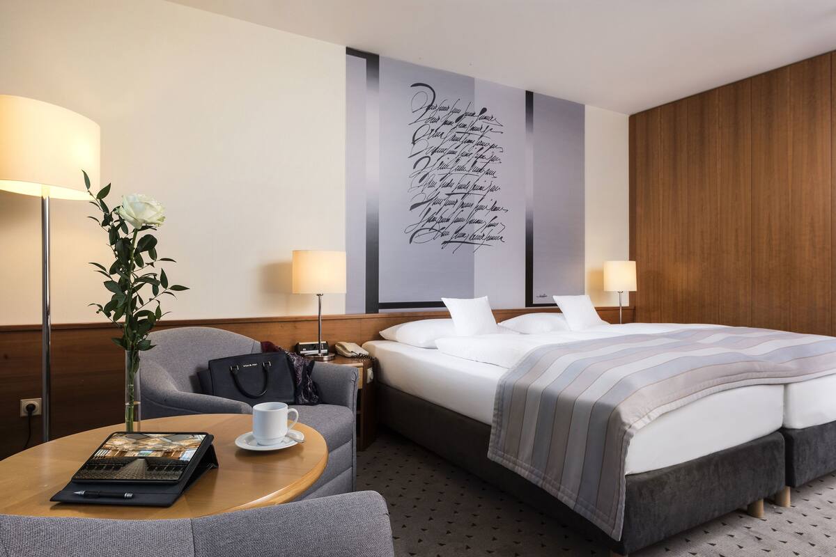 A hotel room with a bed, bedside tables, a desk with a laptop, a chair, a lamp, and a wall decoration.