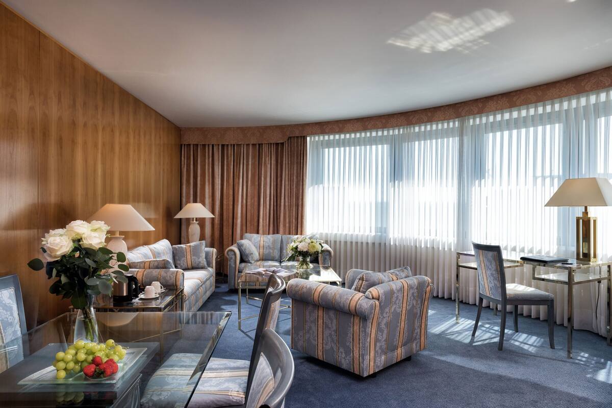 A modern hotel room with a sofa set, a dining table, and a glass coffee table with fruit.