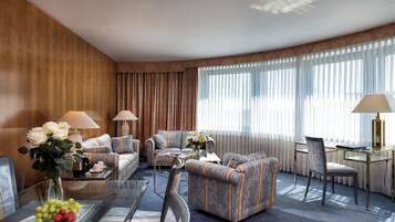 Una habitación de hotel moderna con una pared de acento de madera, un conjunto de sofá, una mesa de comedor con sillas y un ventanal con cortinas traslúcidas.