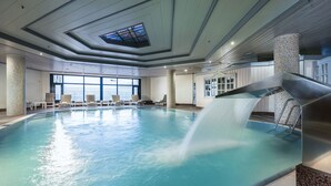 Una piscina interior con tobogán de agua, sillas de descanso y grandes ventanales.