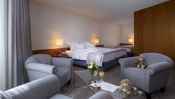A hotel room with two beds, grey upholstered chairs, a glass coffee table with a teddy bear, a vase with flowers, and a glass of orange juice.