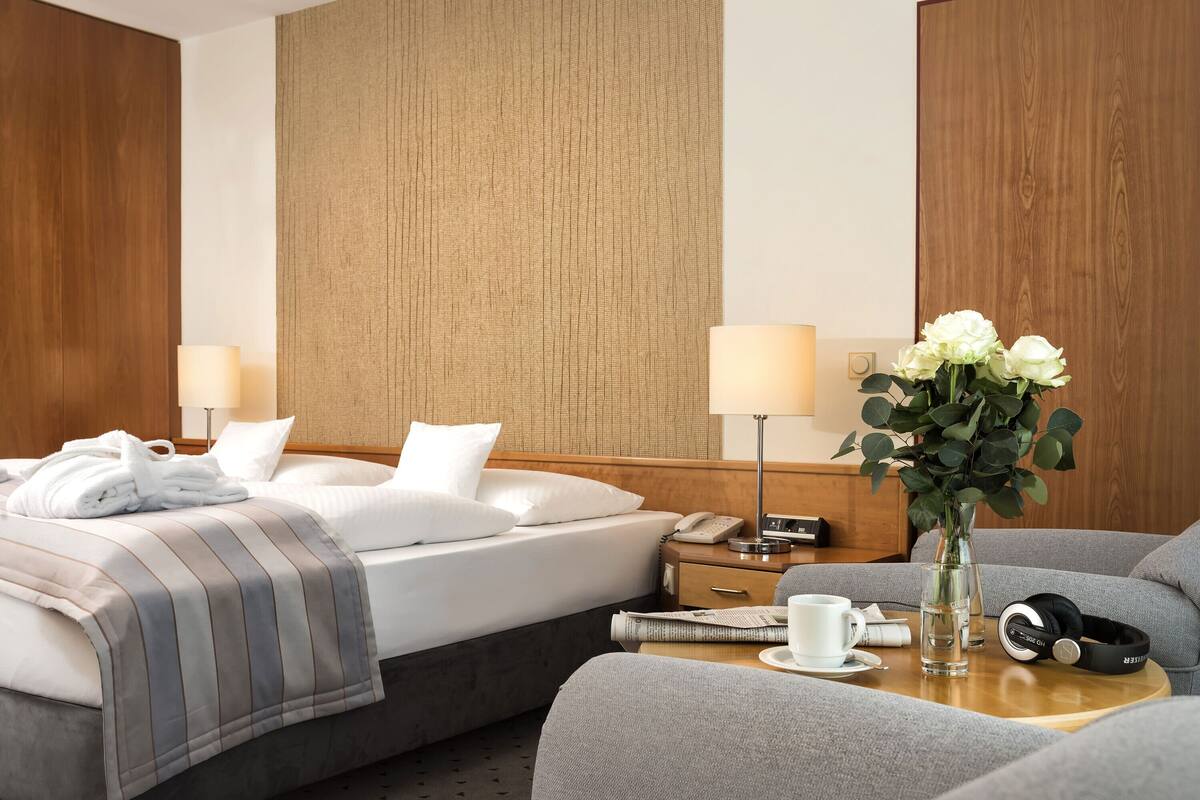 A hotel room with a neatly made bed, two bedside lamps, a vase with flowers, a coffee cup, and a pair of headphones on a side table.