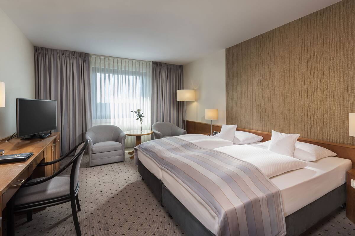 A hotel room with a large bed, a desk with a TV, two chairs, a small table with a vase, and a textured wall.
