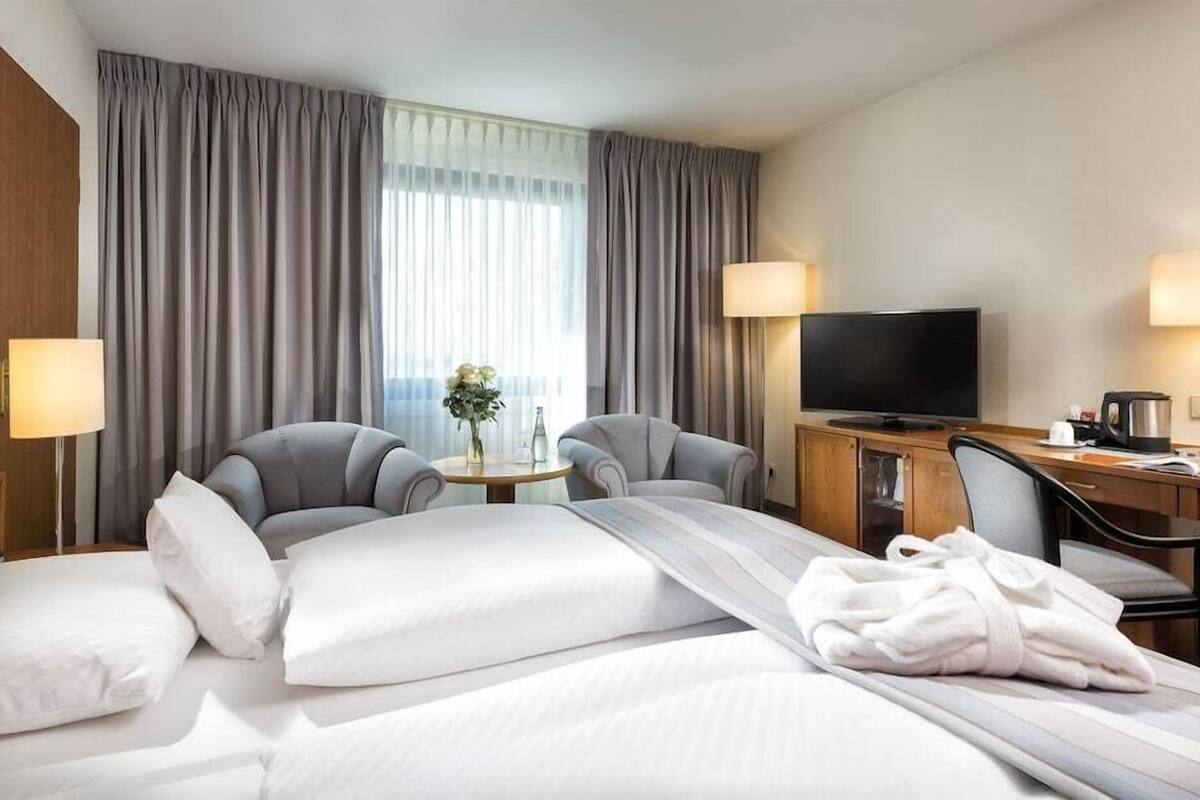A hotel room with a bed, two armchairs, a small table, a TV, and a desk with a lamp.