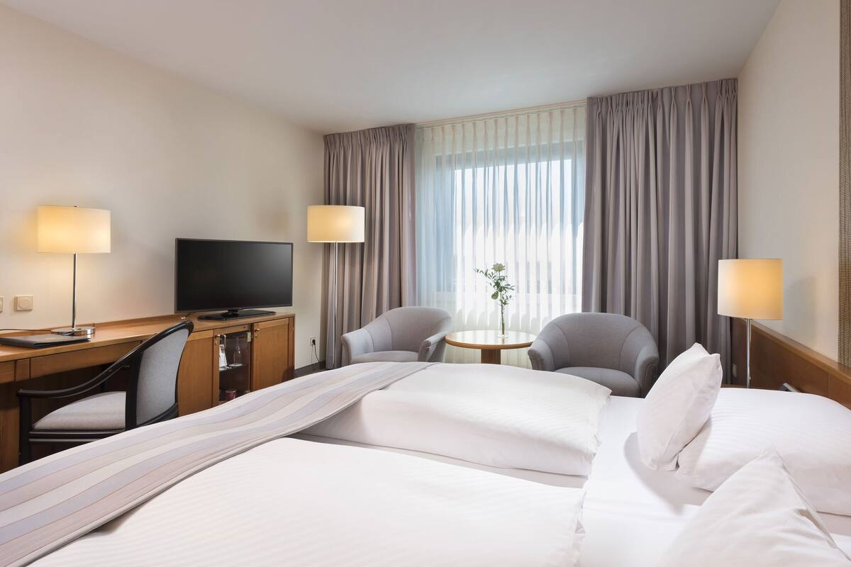 A hotel room with a bed, two armchairs, a desk with a television, and a small table with a plant.