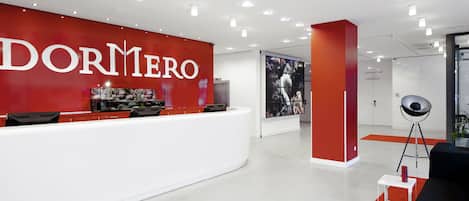 Un área de recepción moderna con una pared roja que muestra 'DORMERO' en letras grandes, un mostrador de recepción con computadoras, un cartel grande y una silla negra con una taza roja sobre una mesa blanca.