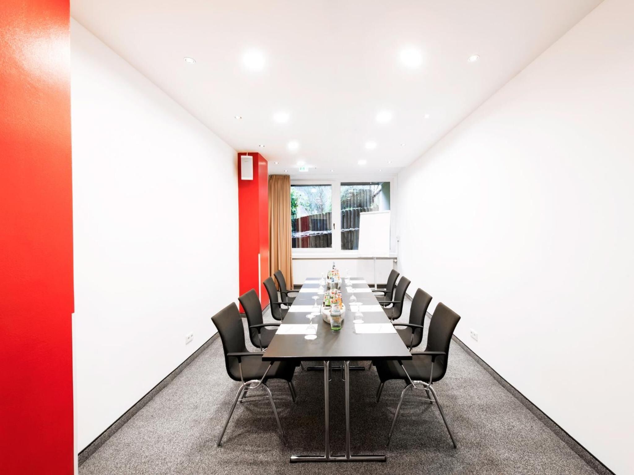 Une salle de conférence avec une longue table, des chaises noires et un mur d’accent rouge.