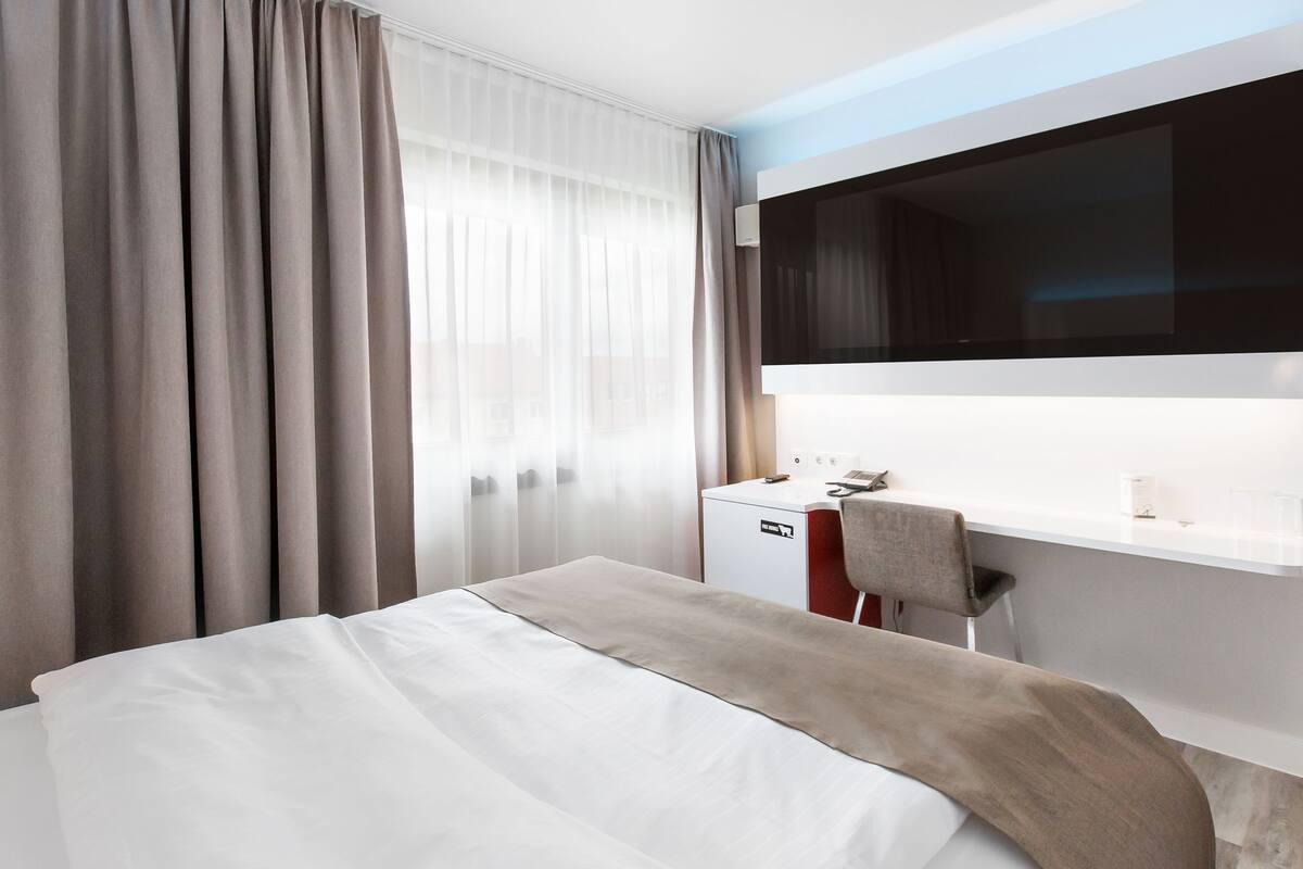 A hotel room with a bed, a desk, a chair, a TV, and a window with curtains.