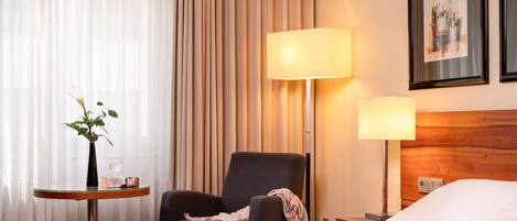 A hotel room with a bed, two chairs, a small table with a vase and a glass, and two wall-mounted lamps.