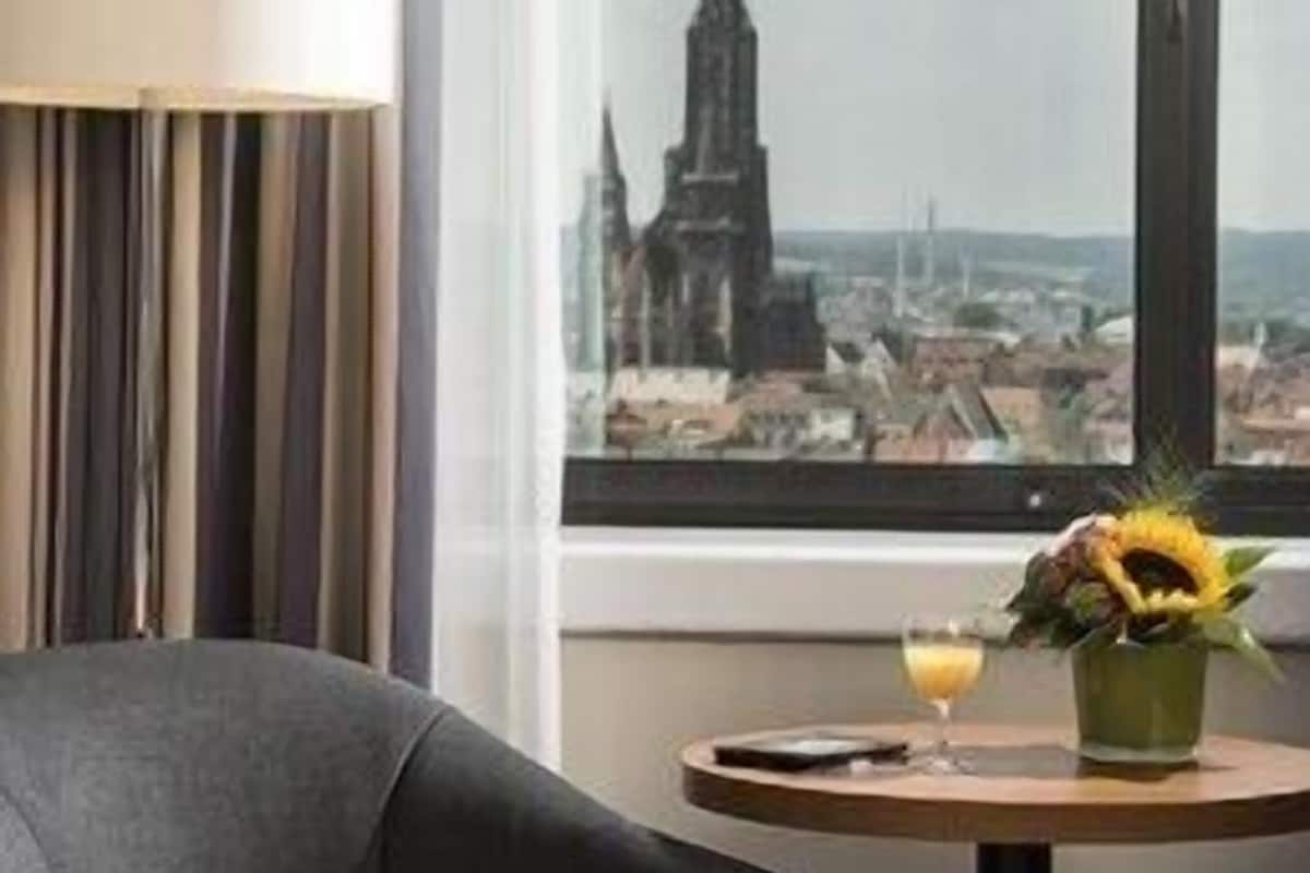 A hotel room with a large window offering a city view, a bed, a table with a drink, and a vase with flowers.