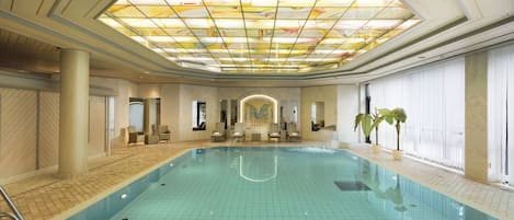 Una amplia piscina cubierta con suelo de baldosas, techo con diseño de vitral y zona de estar con sillas y una mesa.