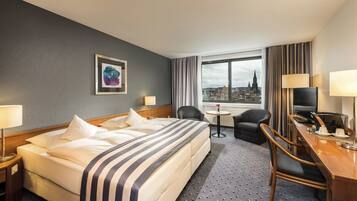 A hotel room with a bed, a desk with a TV, a chair, a small table with a lamp, and a window with a city view.