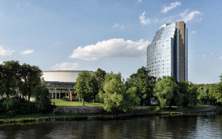 A modern building with a reflective glass facade, a large courtyard with trees, and a body of water in front.