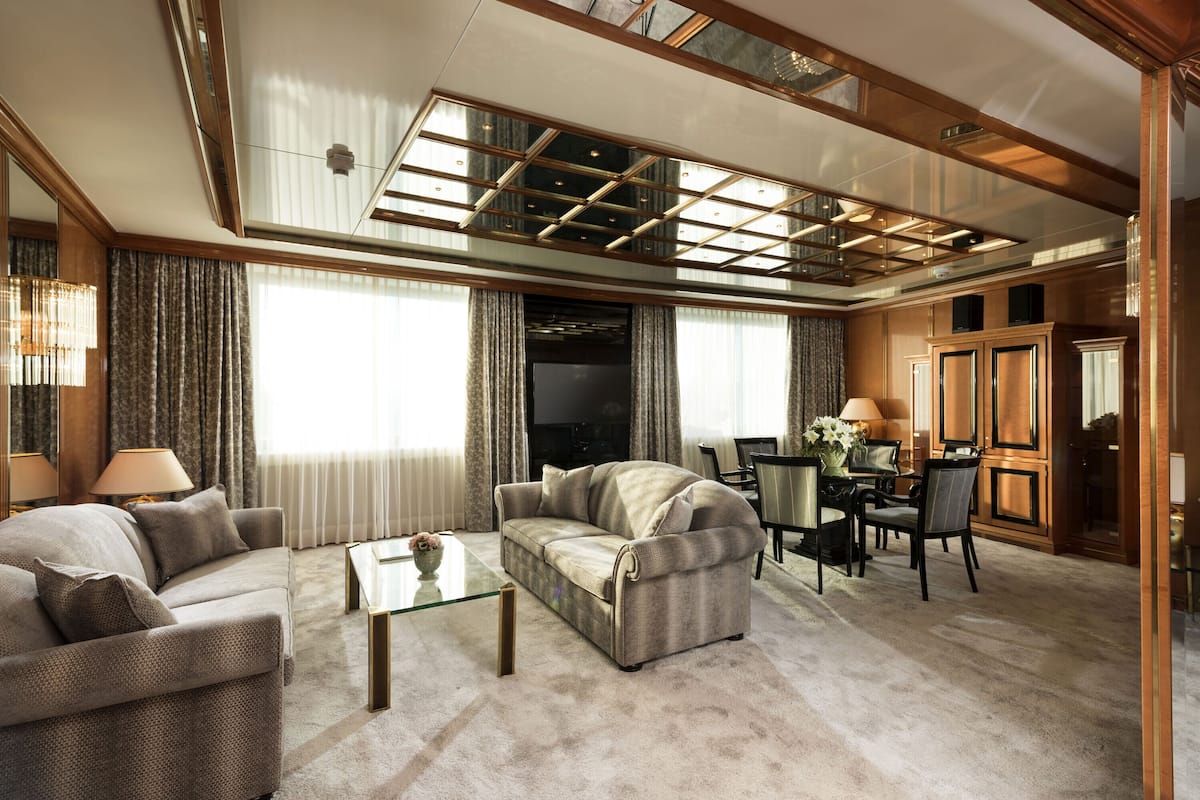 A spacious living room with a large sofa, a coffee table, and a television. The room has a high ceiling with a skylight and wooden paneling.