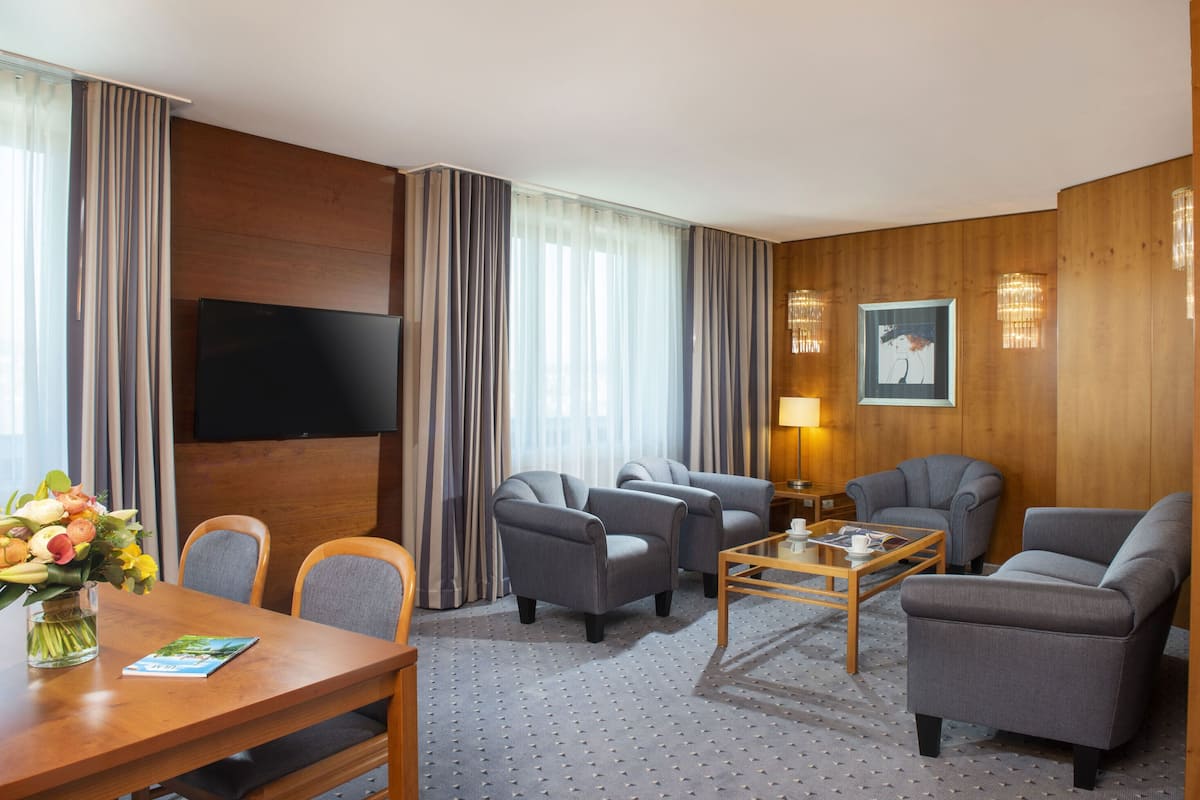 A hotel room with a wooden panel wall, a flat-screen TV, grey upholstered chairs, a wooden coffee table, and a vase with flowers.