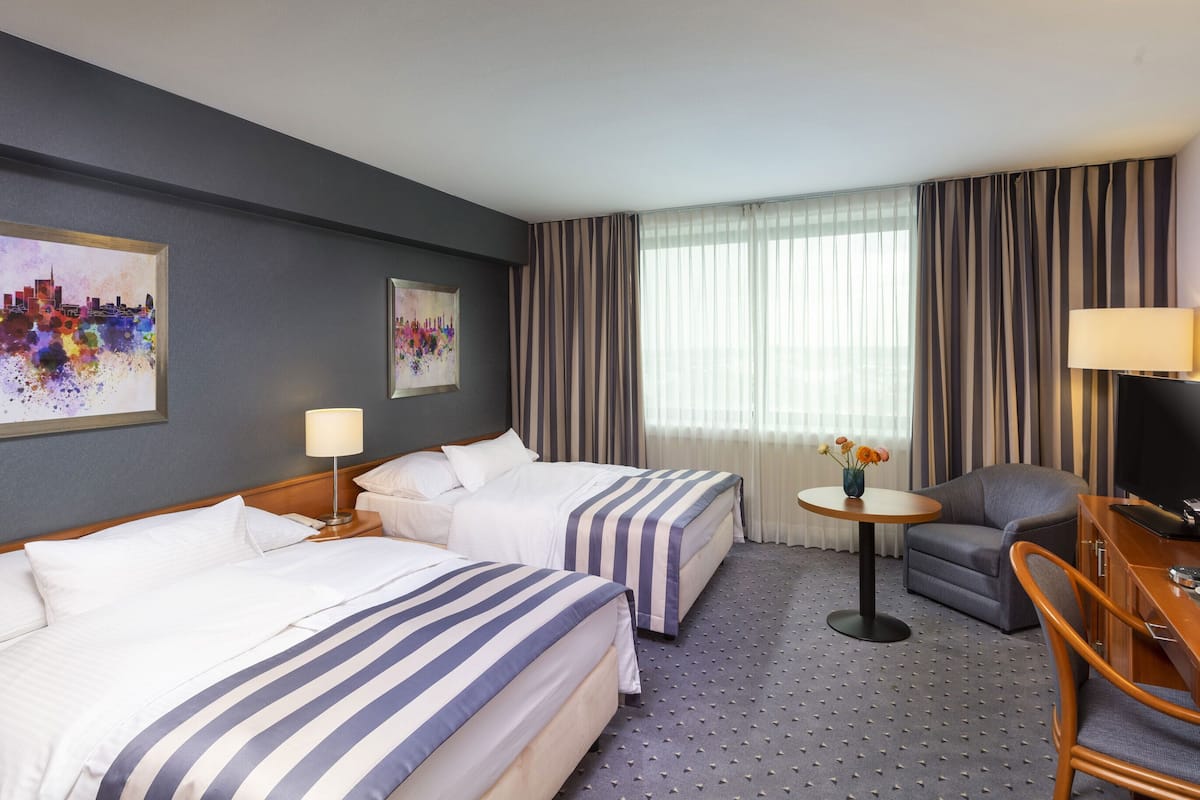 A hotel room with two beds, a desk, a chair, a TV, and artwork on the walls.