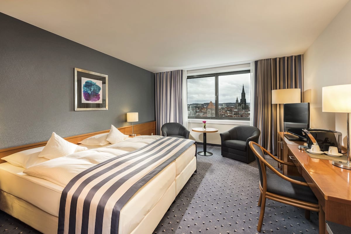 A hotel room with a bed, a desk with a TV, a chair, a small table, and a window with a city view.