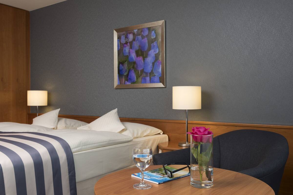A hotel room with a neatly made bed, a bedside table with a glass of water, a book, and a vase with a single rose.