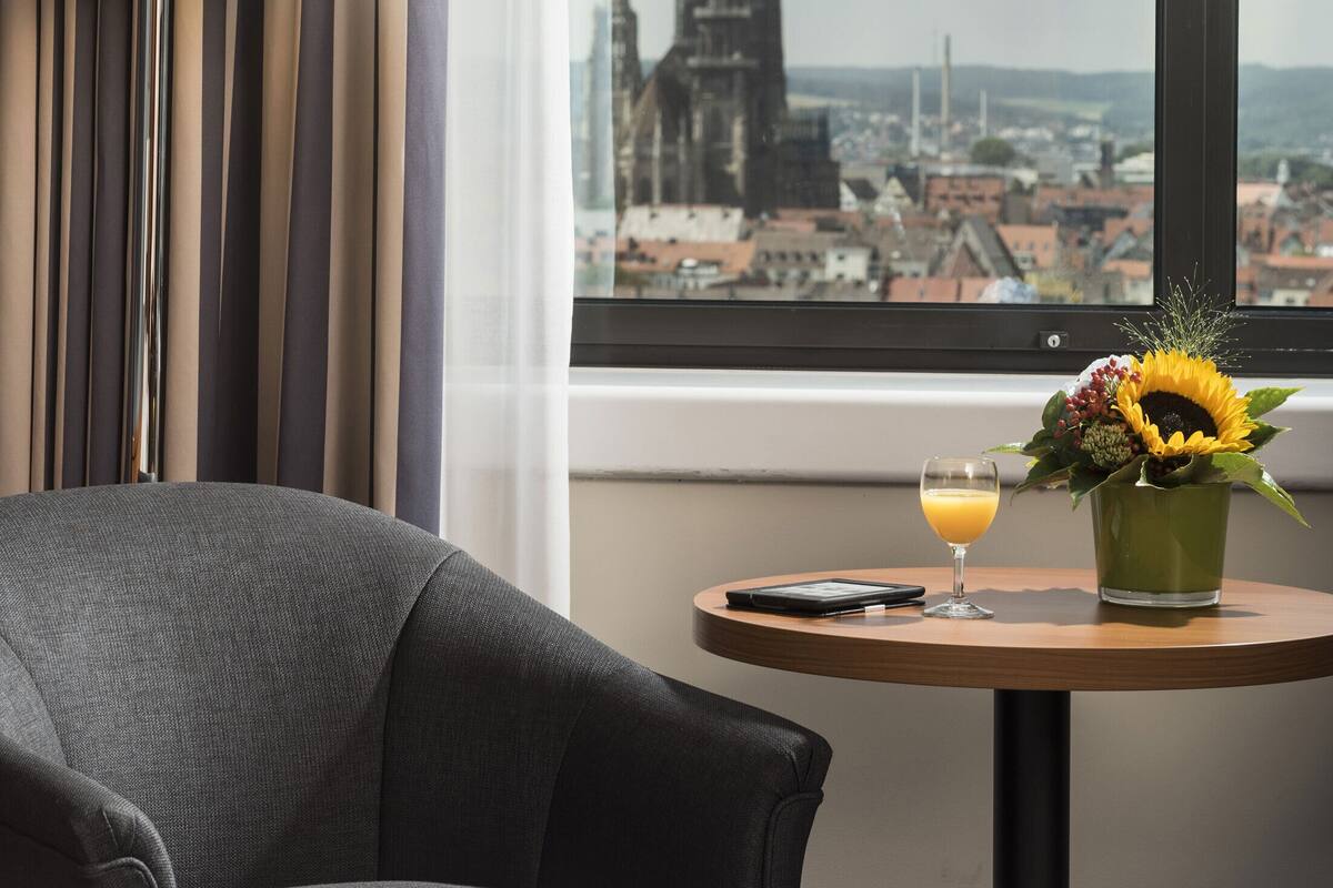 A hotel room with a bed, a lamp, a table with a drink and flowers, and a view of a cityscape through a window.