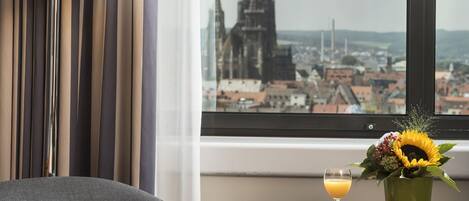 A hotel room with a bed, a table with a drink and flowers, a lamp, and a window with a city view.