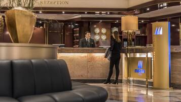 A hotel reception area with a reception desk, a man and a woman standing, and a lounge area with a black leather sofa.